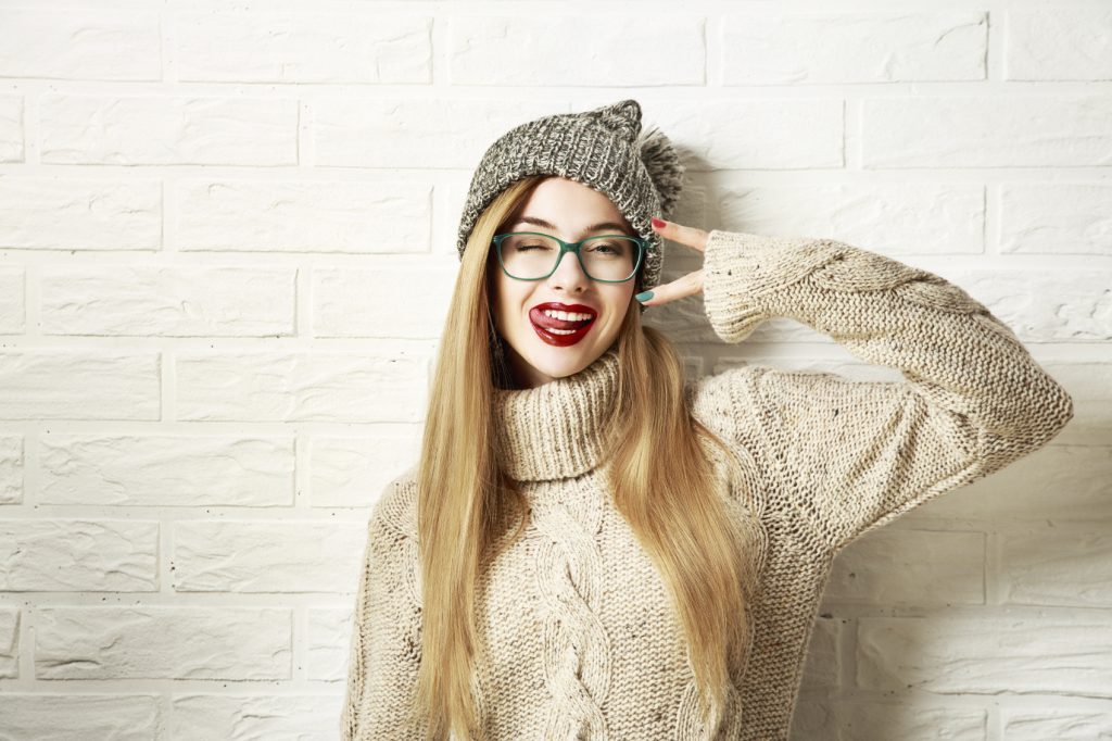 Funny Hipster Girl in Knitted Sweater and Beanie Hat Going Crazy at White Brick Wall Background. Trendy Casual Fashion Outfit in Winter. Toned Photo with Copy Space.